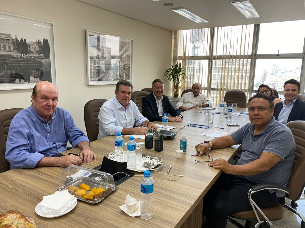 Seis homens e uma mulher sentados frente a uma mesa de reuniões